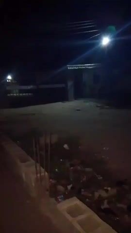 The video captures a dark, outdoor scene at night in Iwerekun, Nigeria. The camera slowly pans across an unpaved area or lot. Bright streetlights are visible, casting light onto the ground, with one particularly prominent light source high in the frame. In the background, a small, simple building is dimly visible. The ground in the foreground and mid-ground is covered with scattered debris and trash. Along the bottom edge of the frame, what appear to be concrete blocks or a low, unfinished wall are present. No individuals or specific activities are shown, only the static nighttime environment. No visible text can be discerned.