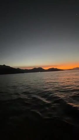 A video shows a body of water under a gradient sky transitioning from dark gray to orange at the horizon. Mountains are silhouetted against the orange glow. The water is dark with visible waves and ripples. A faint crescent moon is visible in the upper left corner of the sky. The overall impression is a twilight or dawn scene at sea.