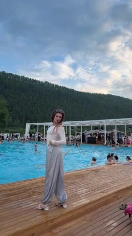 This video captures a vibrant summer day at an outdoor swimming pool complex, likely a resort or entertainment venue, located in Дрокино, Russia. The main subject is a woman with distinctive dreadlocks, striking a pose on a wooden deck overlooking the pool. She is wearing a stylish, off-the-shoulder white top and flowy, light gray pants, paired with white heeled sandals. Her sunglasses are perched atop her head.

The pool is filled with people enjoying the water, with some swimming and others relaxing. Beyond the pool, a canopy structure with white pillars and roofing provides shade, and a DJ booth with equipment suggests music and entertainment are part of the atmosphere. In the background, a lush, tree-covered mountain slopes up towards a partly cloudy, dramatic sky, indicating it is likely late afternoon or early evening. The overall mood appears festive and relaxed, with many individuals engaged in leisure activities.

Several other people are visible, including men walking on the deck. One man in a yellow t-shirt and blue shorts walks past the woman. Another man in a black t-shirt and dark pants walks behind her, and then a man in a yellow shirt and black shorts walks past. These individuals add to the dynamic scene and suggest a social gathering. The wooden deck, polished and well-maintained, forms the foreground, leading the eye towards the pool and the scenic natural backdrop. There is no visible text in the video.
