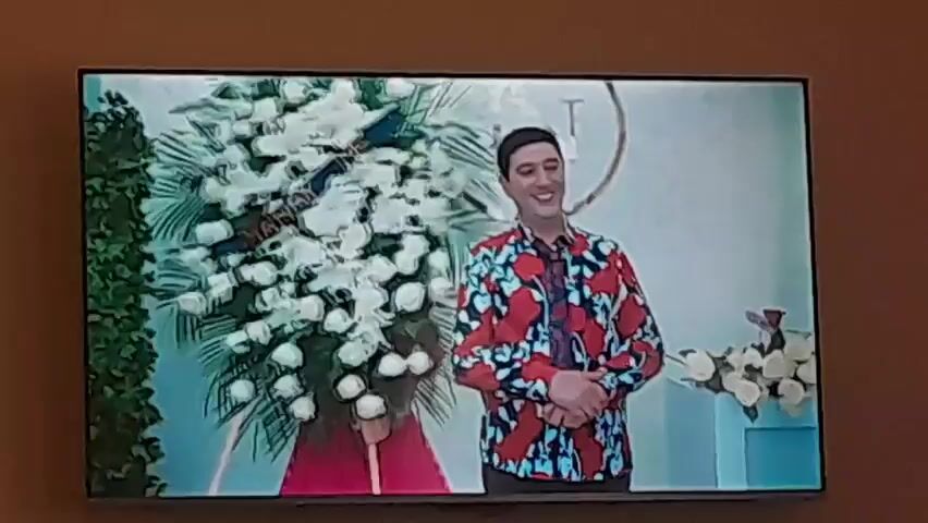 The video displays two alternating indoor scenes. In the first scene, a man with dark hair, wearing a red, blue, and black patterned shirt and dark pants, stands smiling in front of a large white floral arrangement. A woman in a red dress is partially visible behind the flowers, also smiling. The background is a white wall with a circular decoration. In the second scene, a man with a beard wearing a light blue t-shirt stands beside a woman with long dark hair wearing a light yellow shirt. They both look to their left with serious expressions.