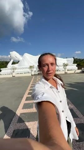 A woman in a white shirt takes a selfie with a reclining Buddha statue in the background. The statue is large and white, with intricate detailing. The woman is smiling and looking at the camera. The sky is blue and there are some clouds. The ground is paved with large square tiles.