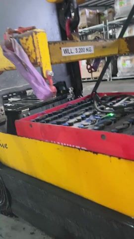 In a storage facility or warehouse environment, a yellow lifting crane is suspended above a large battery bank. The crane's WLL (Working Load Limit) is marked as 3,200 kg. The battery bank is housed in a red and yellow tray or container. Blue and black battery cells are visible, connected by a network of black wires. A green light indicates that the battery is being charged or is functioning correctly. The warehouse contains racks filled with stacked items, possibly containers or boxes. The lighting in the area is industrial, providing bright illumination.