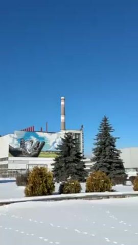 The video shows a large building, likely an industrial or governmental structure, in a snowy, winter setting. The sky is a clear, vibrant blue. The building is light-colored with a tall smokestack near its left side. A large mural depicting hands is painted on the side of the building near the smokestack. Evergreen trees and small, rounded shrubs line the front of the building area. The ground is covered in snow, and there are footprints visible in the snow. The camera pans from left to right, revealing more of the building's facade, including windows and a symbol on another section of the building. There are Ukrainian flags visible near the entrance of the building.