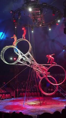 This is a video of a circus performance. There are at least five performers on a complex, metallic structure that resembles a giant pair of scissors. Two performers are standing on top of the structure, each balanced on a large metal ring. Below them, a performer is balancing on the central part of the structure, and two more performers are balanced on lower, larger metal rings. The structure is lit with red and blue lights, and there is an audience seated in bleachers in the background, visible in the upper portion of the frame. The lighting suggests this is an indoor performance, likely in a tent or arena.