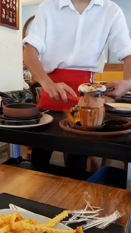 A person wearing a white shirt and a red apron is standing at a table in a restaurant, preparing food. They are using a torch to flambé a dish served in a clay pot. The pot is sitting on a round, dark brown plate, which is on top of a black serving tray. The flames are visible coming from the top of the clay pot, engulfing the food. To the left of the clay pot is another smaller clay pot on a similar plate. In the foreground, a plate of French fries and some toothpicks are visible on a wooden table. The background shows framed pictures on the wall, a clock, and other patrons seated at tables. The lighting appears to be natural and artificial.