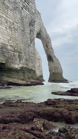 The video shows a coastal scene with towering white cliffs. The cliffs have distinct horizontal striations and one section forms a natural arch. The sea is visible in the foreground, with gentle waves lapping against rocky shorelines covered in dark seaweed. The water appears greenish and somewhat murky. A small archway or cave is visible at the base of the cliff on the left. Two individuals are standing near this opening on the rocky shore. The sky is overcast.