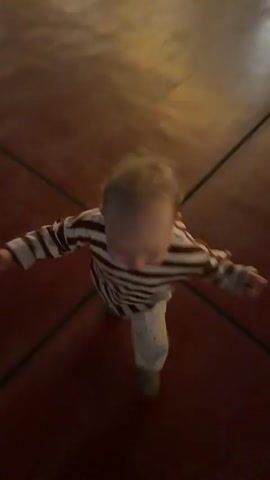 The video shows a person's feet and legs from a first-person perspective. The person is standing on a dark red tiled floor. A baby wearing a striped shirt is being held by the person and then placed on the floor. The baby starts to crawl and then stands up, then falls over. The person's hand is visible patting the baby.