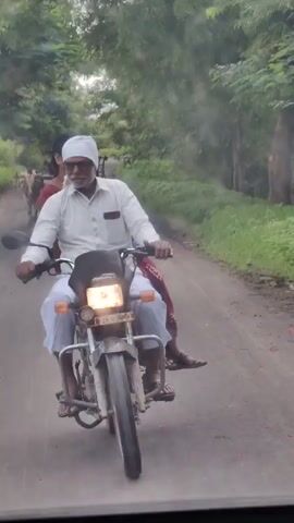 The video shows a man riding a motorcycle on a paved road. The man is wearing a white turban and a white shirt, and he has a beard. He is riding with a person behind him. In the background, there is a bullock cart with two bulls pulling it. The cart is being driven by a man. The road is lined with trees. The scene appears to be in a rural area.