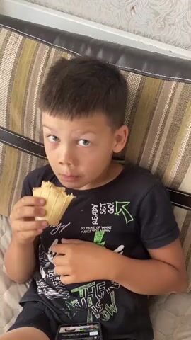 A young boy with short, dark hair and green eyes is sitting on a couch and eating an ice cream cone. He is wearing a black t-shirt with green lettering. The couch has striped pillows. There is a smartphone on the boy's lap. The video is a close-up shot of the boy and his ice cream cone.