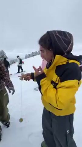 The video shows a young person with long dreadlocks wearing a yellow and black jacket, grey pants, and a dark hood, playing with a Kendama toy on a snowy ski slope. The person successfully swings the ball and catches it on the stick. Another person wearing a plaid shirt and green pants watches them. Other people skiing and snowboarding are visible in the background. The sky is overcast.