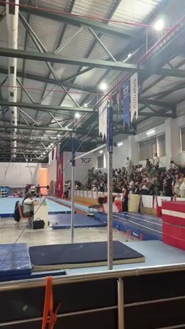 The video shows the interior of a gymnasium during what appears to be a gymnastics competition. In the foreground, there are several blue mats and part of a metal railing. Dominating the center of the frame are the bars of an uneven bars apparatus. Beyond the bars, a blue gymnastics floor is visible, and further back, bleachers filled with spectators are set up. Various gymnastics equipment, including what looks like a vaulting table and other mats, can be seen scattered throughout the gym. Several people are present, including athletes, officials, and spectators. There are flags hanging from the ceiling, one of which appears to be the Turkish flag. The lighting suggests it is daytime, with artificial lights also illuminating the space.