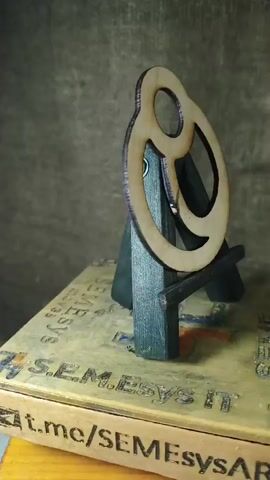 A wooden ornament, resembling a pretzel, is displayed on a small easel. The easel is placed on top of a flat, rectangular object with text and graphics on its surface. The background is a textured, dark gray material. The lighting is soft, with some shadows cast by the ornament and easel. The camera angle is from the side and slightly below the ornament, looking upwards.