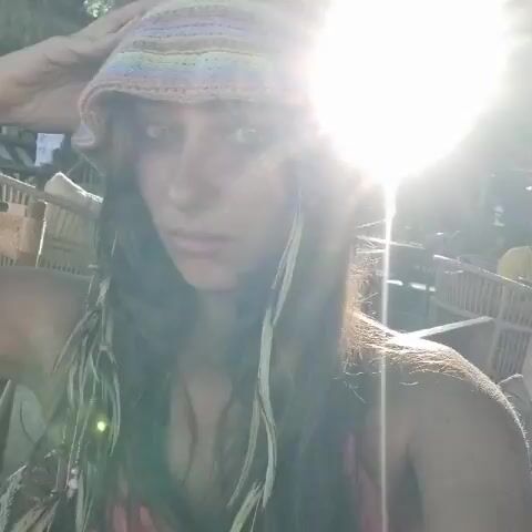 A young woman looks directly at the camera. She wears a crocheted bucket hat with pastel stripes. Her dark hair is shoulder-length, with light-colored hair extensions and beads on either side of her face. She smiles slightly. The background is out of focus but appears to be a tropical outdoor setting with bamboo structures and other people visible. The lighting is bright with some glare.