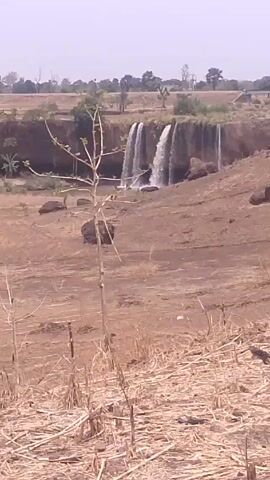 The video shows a waterfall in a dry, grassy landscape. The waterfall has several distinct streams of water cascading over a rocky ledge. The surrounding area is mostly brown and appears arid, with scattered rocks and dry vegetation. In the distance, there are a few trees and what seems to be a structure. The sky is pale and overcast, suggesting it may be a hot day. The camera slightly pans across the landscape.