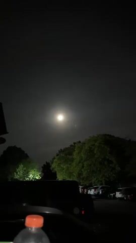The video shows a full moon surrounded by a faint halo in the night sky. The view is partially obscured by trees and cars parked in a lot. A water bottle with an orange cap is visible in the foreground. The video is still and taken at night.