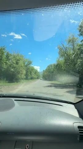 The video shows the view from inside a car, looking out the windshield. The car is driving down a paved road lined with trees on both sides. The sky is blue with some white clouds. In the distance, there are mountains. The road appears to be in good condition, with some cracks and potholes. There is another car visible in the distance, ahead of the camera. The interior of the car, specifically the dashboard, is visible in the foreground. The dashboard is gray and appears somewhat dusty. There are some smudges and marks on the windshield, as well as some debris on the dashboard. The overall impression is of a sunny day driving through a rural or natural landscape.