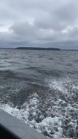 A video shows water splashing from a boat on a cloudy day. The boat appears to be moving, as indicated by the water disturbance at the side of the frame. In the distance, there is a body of water with a landmass visible on the horizon. The sky is overcast with dark clouds. Some fishing rods are visible at the bottom left of the frame.