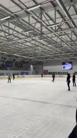 The video shows a wide, brightly-lit ice rink with multiple ice skaters practicing. The rink is enclosed in a modern building with a high ceiling supported by a network of steel beams and with large overhead lights. There are several skaters distributed across the rink, most wearing dark clothing. One skater is wearing a striped shirt and black pants. In the background, there is a banner with the word "ISRAEL" and another with the words "GearGuard". A digital screen displays an image and the letters "OSN". The camera slowly moves around the rink to follow the skaters. The skaters are performing various movements, indicating they are practicing figure skating.