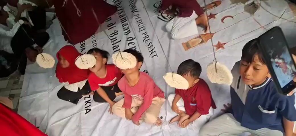 The video shows a group of children sitting on a white tarp, participating in a cracker-eating contest. Each child has a cracker hanging from a string in front of their face, and they are attempting to eat the cracker without using their hands. The children are dressed in casual clothing. In the background, there are adults and other children, and a banner with text in Indonesian. The overall setting appears to be an outdoor or semi-outdoor event.