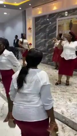 In a bright, spacious room with white walls, a group of women in matching white blouses and maroon skirts dance energetically. Other people are present, some also dancing, and a few dressed more formally. The room appears to be a gathering space, possibly a church or a large hall, with a prominent poster on the wall. Overhead lighting is modern, with blue and yellow accents. The floor is light-colored and shiny, reflecting the light. People are smiling and moving with enthusiasm.