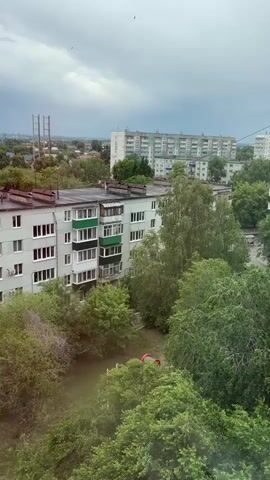 The video presents a static, elevated view of a residential area in Zhemkovka, Russia, on an overcast day. Dense green trees and bushes fill the foreground and much of the mid-ground, interspersed with buildings. A multi-story apartment building with light grey facades and green-framed balconies is prominent in the mid-ground. In the background, several other large, multi-story apartment blocks of similar architectural style stretch across the horizon. Further in the distance, industrial-looking structures with tall towers are faintly visible against the sky. The sky is completely cloudy and grey, indicating an overcast condition. A thin wire crosses the upper right portion of the frame. No people or specific activities are depicted, and no text is visible within the scene.