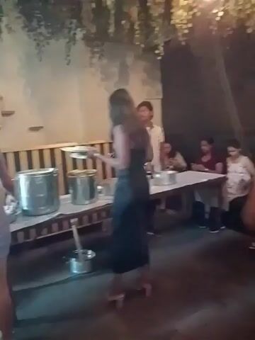 Several women are gathered at a buffet-style meal in an outdoor-indoor area. The space is decorated with hanging plants and warm lighting.  A long white table holds various food items in stainless steel containers and plates. The women are dressed in casual dresses of various lengths and colors. They are seen picking up plates, serving themselves food, and conversing with each other. Men are also seated at a nearby table.