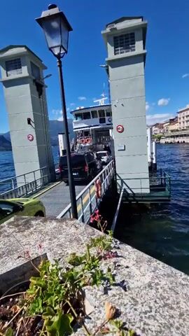 A ferry is docked at a terminal, with two cars lined up on its deck. A yellow hatchback is parked on the left side of the ramp, and a black van is driving onto the ferry from the right. The ferry has multiple levels and appears to be carrying passengers. Two tall, grey towers stand on either side of the ramp, and a lamppost is positioned in the foreground. The scene is set on a body of water under a clear blue sky, with buildings and greenery visible on the distant shore. The water is calm, and the overall atmosphere is sunny and serene.