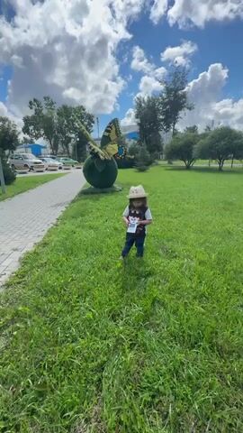 A child, wearing a straw hat, a t-shirt featuring Batman, and jeans, stands on a grassy lawn, holding a toy. Behind the child, a large, green, spherical sculpture adorned with a yellow and black butterfly is visible. The butterfly's wings are spread, and it appears to be perched on the sphere. The scene is set outdoors, with lush green grass and trees in the background. The sky is bright blue with fluffy white clouds, suggesting a sunny day. In the distance, a paved pathway and several parked cars, including taxis, are visible. The overall mood is cheerful, with the child smiling. The time of day appears to be daytime, possibly late morning or early afternoon, given the bright sunlight. The location is identified as Annino, Russia.