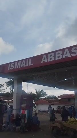 A gas station canopy with the name ISMAIL ABBAS NIGERIA LTD is visible. Several people are gathered underneath the canopy and in the surrounding area. Some individuals appear to be vendors or customers, with some seated and others standing. Various vehicles, including a white pickup truck and a white van, are parked in the background. The ground appears to be paved. The sky is cloudy. The overall scene suggests a busy public space, possibly a marketplace or transportation hub associated with the gas station.