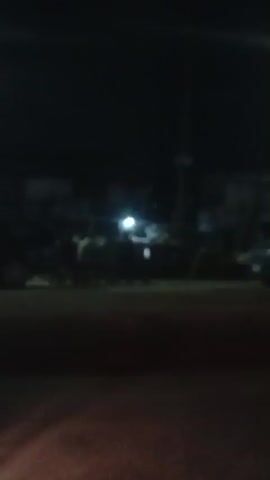 The video appears to be taken at night. The lighting is dim, and the scene is somewhat blurry. In the background, there is a group of people standing near a street, with some vehicles present. There is also a tall structure that looks like a light pole or a tower. The foreground is dark and out of focus.