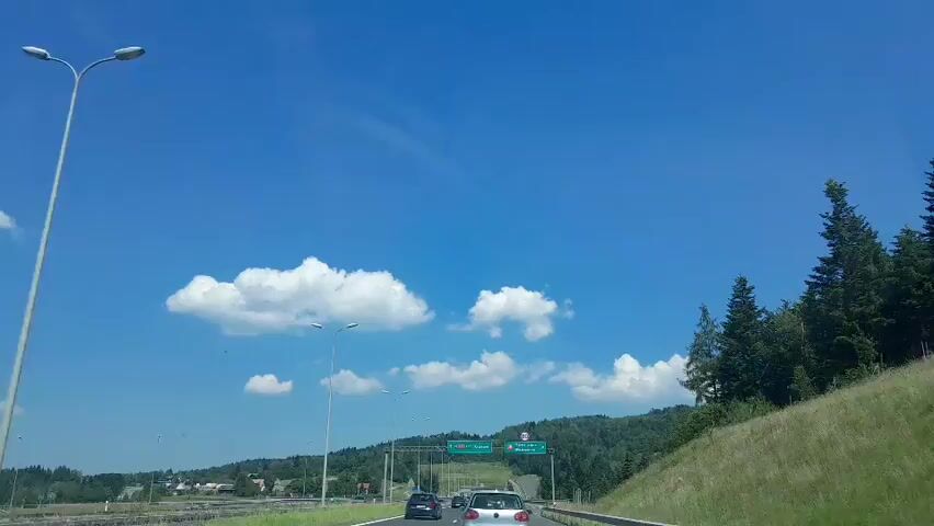 The video shows a view from a vehicle traveling on a road on a clear, sunny day. The sky is bright blue with scattered white clouds. On the right side of the road, there is a grassy hill with trees. In the distance, there are more trees covering rolling hills. On the left side of the road, there are some buildings and more trees. There are streetlights visible along the road. A highway sign is visible overhead, indicating directions to Krakow and Nowy Sacz/Wadowice, with a speed limit sign of 80. Several cars are driving on the road ahead, and the vehicle from which the video is filmed appears to be in motion, moving forward.