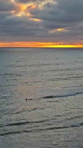 The video shows a view of the ocean at sunset. The sun is setting on the horizon, casting a warm orange glow across the sky. Clouds are present in the sky, obscuring some of the sun. Two people are visible in the water, appearing to be surfers. The waves are small.