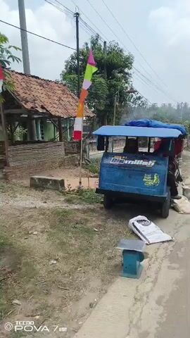 The video shows an outdoor scene with a small blue motorized tricycle parked on a dirt and gravel surface. The tricycle has a blue canopy and is decorated with text and images. In the background, there is a rustic wooden structure with a tiled roof and some vegetation. A colorful flag is visible on a pole near the tricycle. A person, wearing a blue hooded jacket, is partially visible on the right side of the frame, near the tricycle's driver's seat. A large white bag with some print on it is on the ground in front of the tricycle, and a small scale is also present. The video pans to show the side and rear of the tricycle, which has a red and green machinery attachment at the back, possibly for agricultural processing. More bags and debris are scattered around the area. The overall environment appears to be rural.