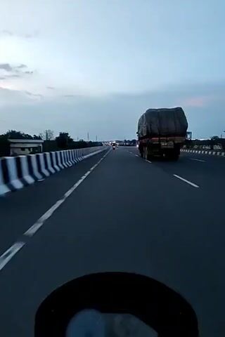 A first-person perspective video shows a motorcyclist driving on a highway. The sky is overcast with clouds, indicating either early morning or late evening. On the left side of the highway, a concrete barrier with black and white stripes is visible. In the distance, several vehicles, including a truck and a car, are traveling in the same direction. On the right side of the highway, buildings and trees can be seen. The road appears to be well-maintained, with clear lane markings.