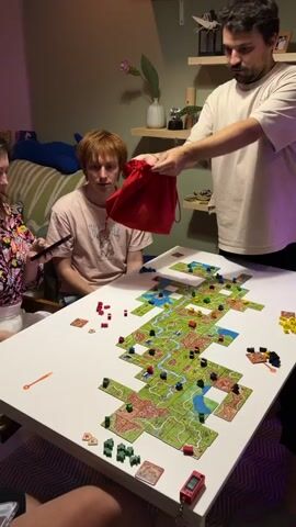 A group of three people are gathered around a large white table playing a board game. The game board, composed of many interlocking tiles, covers most of the table's surface and depicts a landscape with fields, roads, cities, and rivers. The players are using various colored wooden pieces, likely meeples or tokens, to mark their territories and progress in the game.

In the foreground, a woman with dark hair, wearing a colorful floral-patterned top, is reaching towards a red drawstring bag held by a man standing to her right. She appears to be looking at a mobile phone in her left hand. The man holding the bag has dark hair and a beard, and is wearing a light-colored t-shirt. He is holding the red bag in his outstretched right hand, and his left hand is on the bag.

In the middle ground, a young man with reddish-brown hair, wearing a light-colored t-shirt with a graphic design, is seated and looking at the board game. His hands are resting on the table in front of him.

The environment appears to be a room, possibly a living room or dining area, with a striped sofa visible in the background to the left. There are shelves on the wall behind the standing man, adorned with various items, including what looks like a plant and decorative objects. The lighting in the room seems to be a mix of natural light and artificial lighting, with a pinkish hue noticeable on the left side of the frame, possibly from a lamp. The overall atmosphere suggests a casual gathering focused on playing a board game.