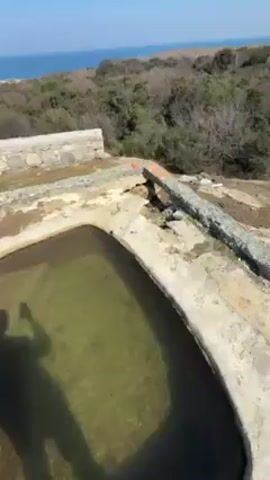 The video shows a concrete structure with a circular pool of water on top of it. The structure is surrounded by scrubby vegetation and the ocean is visible in the distance. The sky is blue and clear. The camera pans across the scene slowly.