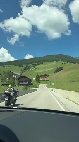 The video shows a view from inside a vehicle driving on a paved road through a scenic mountain landscape on a sunny day. The road curves to the right, with a motorcycle visible in front of the vehicle. The landscape is characterized by rolling green hills dotted with farmhouses and trees, and the sky is blue with scattered white clouds. There are barriers along the side of the road. A sign with an image of a bus is visible on the right side of the road. The word "BUS" is painted on the road in yellow. The scene appears to be in a rural, mountainous region.