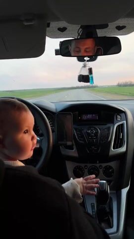 A baby is seated in the front passenger seat of a car, facing towards the dashboard and the road ahead. The baby has light hair and is wearing a light-colored top under a dark garment, possibly a car seat strap or part of their clothing. Their left hand is near the gear stick, and their right hand is near the dashboard.

The car's interior is visible, including the steering wheel, a dark-colored dashboard, an infotainment system, and air vents. A smartphone is mounted vertically on the dashboard, displaying an image or video. In the rearview mirror, the reflection of an adult driver is visible, looking towards the road. A small dashcam and a dangling air freshener (a blue liquid vial and a black bag) are also attached to the rearview mirror.

Through the windshield, a paved road stretches into the distance, flanked by green fields and distant trees. The sky above is soft with hues of pink and blue, suggesting late afternoon or early evening. The car appears to be in motion on a highway or a major road. Visible text includes "FOCUS" on the dashboard and "MUSIC" on the infotainment screen. The scene takes place in Vyselki, Russia.
