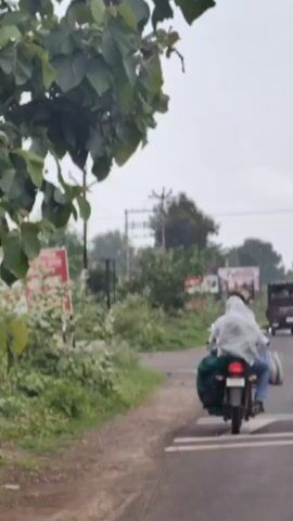 The video shows a motorcyclist and a passenger riding a motorcycle on a paved road. Both individuals are covered by a light-colored sheet, presumably to protect them from the elements. The motorcycle is carrying several bags, including a large green bag on the back and a striped bag on the side. The road is flanked by greenery, and there are utility poles and wires visible in the background. In the distance, other motorcycles and a truck can be seen. A billboard with the text "Fulltoss" is also visible on the side of the road. The sky is overcast.