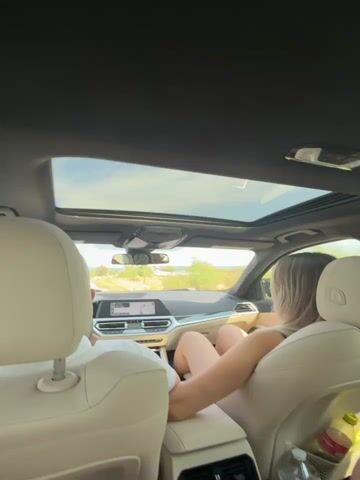 This video shows the interior of a car with a panoramic sunroof. Two individuals are visible. One person, facing away from the camera, is in the passenger seat of the front row, with their legs extended. Another person is in the back seat, also facing away from the camera. The car appears to be in motion, with the exterior landscape visible through the windows and sunroof. Several objects are present within the car, including water bottles and a bag. The lighting suggests it is daytime.