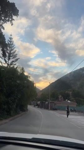 The video shows a view from inside a car driving on a road toward a mountain at sunset. The road has a white line down the center, and there are trees and buildings on either side of the road.  Several people are walking on the side of the road. There are power lines running overhead. The sky is a mix of blue and gray clouds, with the sun setting behind the mountain.
