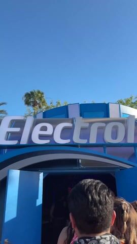 The video captures a bright, sunny outdoor scene at what appears to be a music festival or large event in Indio, United States of America.

The primary subject is a large, futuristic-looking blue and white structure with the word "Electro" prominently displayed in large white letters across its arching facade. This structure serves as an entrance, with several people queuing and walking into a darkened interior. Above the structure, a clear blue sky is visible, along with the tops of palm trees and other green foliage in the background.

At the 0:02 mark, the camera focuses on a young woman with reddish-brown hair, wearing a white top and a white headscarf. She smiles at the camera, expressing excitement by saying "So excited!" She is holding a white can, which appears to be a White Claw Hard Seltzer, with "140 CALORIES" and "2g CARBS" visible on the label. She then makes a playful, slightly disgusted face while speaking, mentioning "...un shot con en el..." (a shot with in the...), indicating she is discussing the drink. The background remains the "Electro" entrance, with other attendees visible in the queue.

Upbeat electronic music is audible throughout the clip, adding to the festive atmosphere. The bright daylight, clear sky, and presence of palm trees suggest a warm climate and an outdoor event setting.