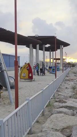 A vertical video shows an outdoor playground with various structures and people, with a long white metal fence on the right separating the playground from a rocky, natural area. The sky is partially cloudy, and the sun, setting in the distance, casts a golden glow on the horizon. The playground features slides, climbing structures, and shaded areas with wooden roofs supported by pillars. Several adults and children are visible on the playground, some near the play equipment and others seated under the shaded structures. The rocky area on the right appears to be a natural barrier or coastline, with sparse vegetation growing among the stones. In the background, buildings and distant hills are faintly visible under the colorful sky. The video slowly pans to the right, revealing more of the playground and the distant sunset.