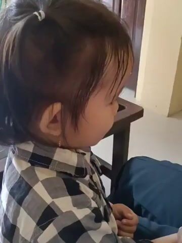 A child wearing a black and white checkered shirt is being fed a small green object from a spoon by an unseen person. The child is facing to the right and looking down at the spoon. The background appears to be an indoor setting with wooden furniture and walls.