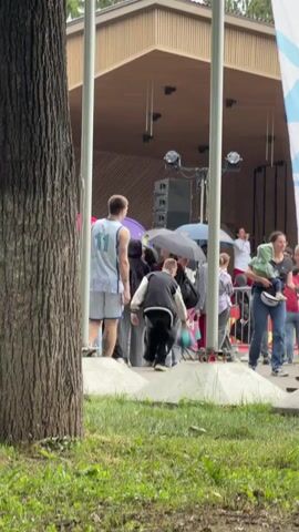 This is a medium shot taken outdoors on a cloudy day, possibly in the late morning or early afternoon. The scene is set in a park or open area with a stage in the background and a large tree trunk in the foreground. A group of people are gathered around, some holding umbrellas, suggesting it might be raining or threatening to rain.

The central subject is a young man wearing a blue and white patterned athletic tank top with the number "11" on it, and matching shorts. He is facing away from the camera and appears to be moving or dancing. To his left, a young woman in a black bomber jacket and black pants is also moving. Several other people are visible in the background, some holding umbrellas. A child is perched on someone's shoulders in the distance.

The stage in the background has a wooden canopy and appears to be set up for a performance, with speakers and lighting equipment visible. A large white and blue banner is partially visible on the right side of the frame. The ground is covered in grass and a concrete barrier separates the grassy area from where the people are gathered.

There is no visible text in the image. The overall atmosphere seems to be one of a casual outdoor gathering or event.