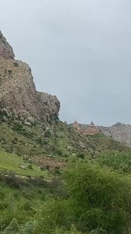 A daytime shot of a monastery on a hill. There are two dome structures visible on the monastery. The hill is covered in green shrubs and trees, and there are some large rock formations to the left of the monastery. The sky is overcast and grey.