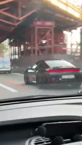 The video shows a dark-colored Porsche coupe seen from inside another vehicle, apparently on a road. The Porsche is in front, moving in the same direction. Initially, the vehicles are under a bridge-like structure with red supports. Later, they are on an open road with a pedestrian overpass and road signs visible. The Porsche has a light bar-style taillight design and a Swiss license plate. A white van is also visible in the periphery. The footage is taken from the driver's seat of the trailing car.