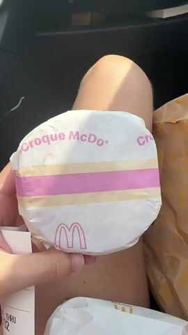 A person's hand holds a round, white McDonald's burger wrapper with pink and yellow stripes. The words "Croque McDo" are printed in pink at the top of the wrapper, and the McDonald's logo is visible in pink near the bottom. The wrapper is held in front of the person's knee and thigh, which are visible. A receipt is also partially visible in the lower left corner. In the background, a brown paper McDonald's bag is visible. The scene appears to be inside a car, suggested by the dark interior visible in the upper left corner. The lighting indicates it is daytime.