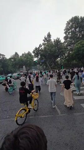 A group of pedestrians and cyclists are crossing a busy street with multiple lanes of traffic. The scene appears to be a city street with trees lining the sides and traffic lights visible in the distance. Some people are walking, while others are riding bicycles, including one person on a yellow bike. The sky is overcast.