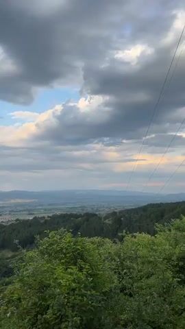 The video shows a panoramic view of a valley with a town nestled within it, surrounded by rolling hills covered in lush green trees. The sky is filled with dramatic clouds, some dark and heavy, others lighter and tinged with the soft light of dawn or dusk. The camera pans slowly across the landscape, capturing the vastness of the valley and the distant mountains. The overall impression is one of natural beauty and tranquility, with the town appearing as a small, human element within the grand scheme of nature. There are no people or specific objects prominently featured in the video, focusing instead on the atmospheric landscape.